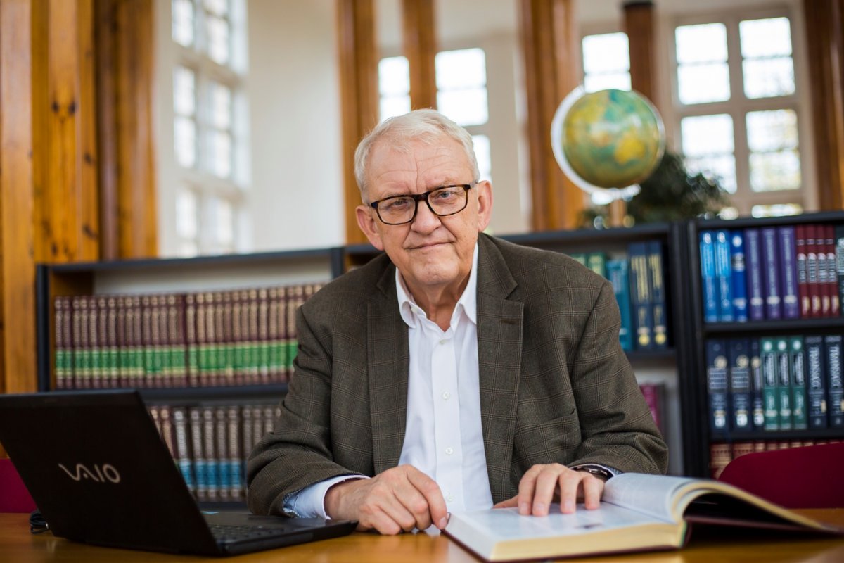 Prof. Juliusz Gardawski - portrait photo on the background of bookshelves. In front of the Professor there is a laptop and an open book on the table