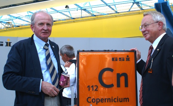 Zbigniew Majka i Sigurd Hofman na uroczystości poświęconej odkryciu pierwiastka Copernicium, pozują przy dużym pomarańczowym symbolu pierwiastka