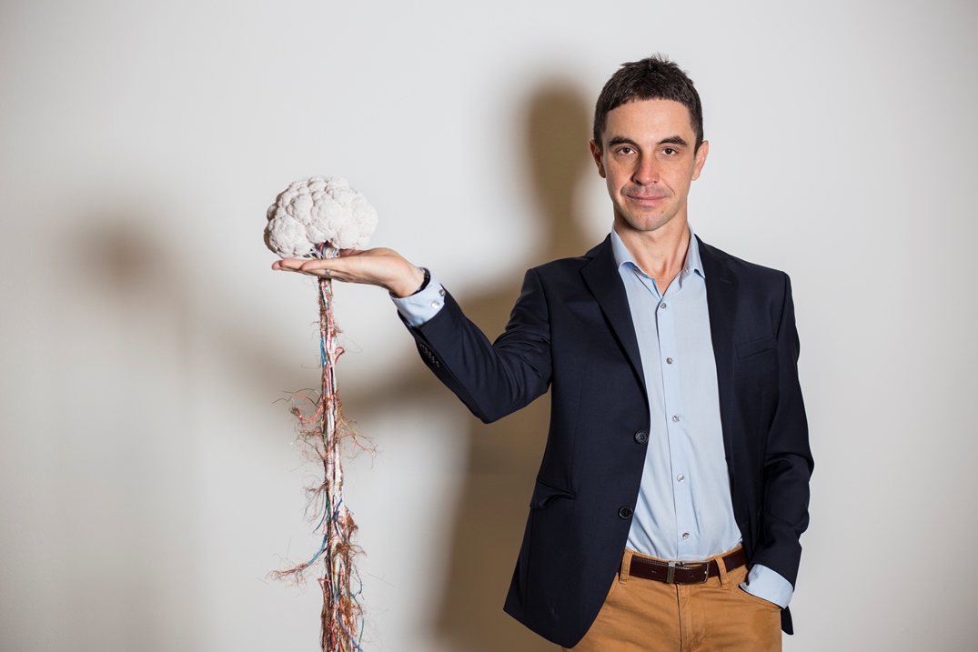 Marcin Szwed pozuje obok rzeźby przedstawiającej ludzki mózg umieszczony na pionowym wąskim postumencie splecionym z drutów.